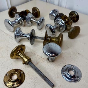 May include: A collection of vintage door knobs in various styles and finishes. The knobs include polished chrome, brass, and aged bronze. Some have oval shapes, while others are round. The hardware is arranged on a light-colored surface.