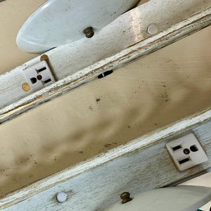 May include: Close-up of a section of a vintage light fixture. The fixture has a distressed, off-white and light blue painted wooden frame. White electrical outlets are visible, along with white, oval-shaped light covers and brass-colored hardware.