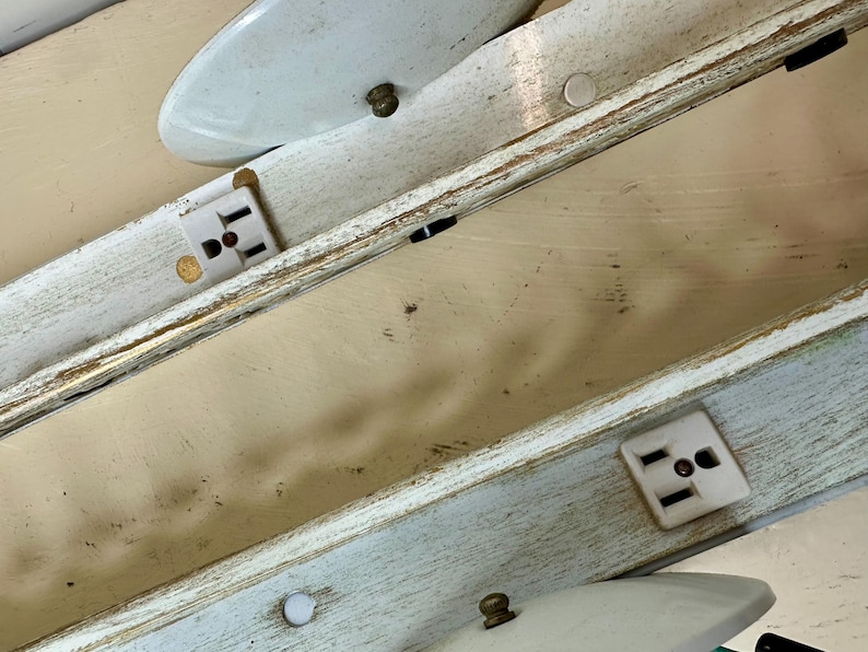 May include: Close-up of a section of a vintage light fixture. The fixture has a distressed, off-white and light blue painted wooden frame. White electrical outlets are visible, along with white, oval-shaped light covers and brass-colored hardware.