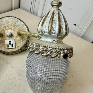 May include: An antique wall sconce with a clear, textured glass shade and ornate, off-white metal detailing. The sconce features a decorative top and a circular backplate with a power outlet. The metal has gold accents and a distressed finish.