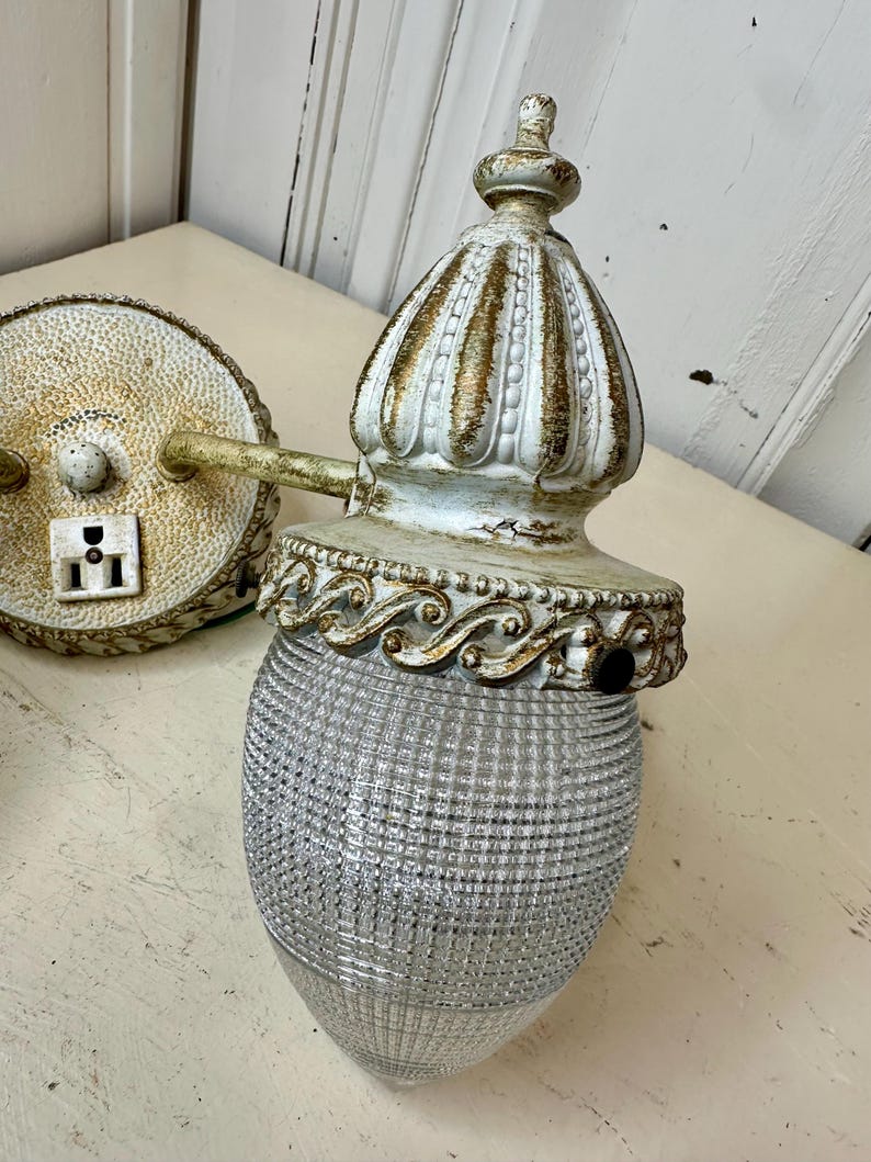 May include: An antique wall sconce with a clear, textured glass shade and ornate, off-white metal detailing. The sconce features a decorative top and a circular backplate with a power outlet. The metal has gold accents and a distressed finish.