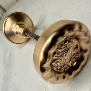 May include: A vintage-style door knob with a warm, metallic finish. The knob features an oval-shaped design with an ornate, embossed floral pattern. The hardware includes a round base and a connecting rod.