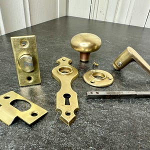 May include: A collection of antique brass door hardware components, including a doorknob, lever handle, escutcheons, and a bolt. The hardware has a warm, golden hue and shows signs of age.