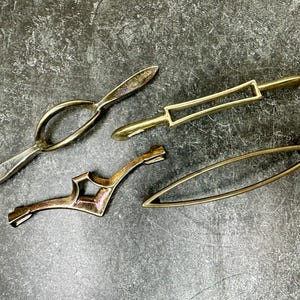 May include: Four antique brass hardware pieces are arranged on a dark, textured surface. The pieces include a curved handle, a rectangular handle, a decorative bracket, and an oval-shaped pull.