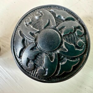 May include: A black metal knob with a decorative floral design. The knob has a raised center and a textured surface.