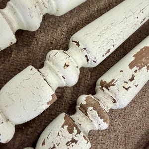 May include: Three wooden furniture legs featuring a turned, spindle-style design. The legs are finished in a distressed, off-white paint that is cracked and peeling in various areas, revealing the natural brown wood grain underneath the surface.