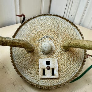 May include: An antique, round, off-white electrical fixture with a textured surface and a decorative, scalloped edge. It features a white outlet, two curved arms, and exposed wiring. The fixture has a vintage, aged appearance.