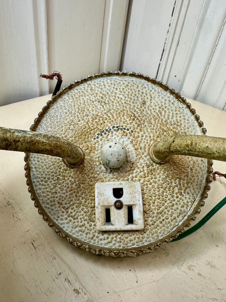 May include: An antique, round, off-white electrical fixture with a textured surface and a decorative, scalloped edge. It features a white outlet, two curved arms, and exposed wiring. The fixture has a vintage, aged appearance.