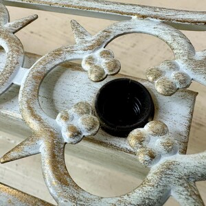 May include: Close-up of a decorative metal fixture with a distressed white and gold finish. The design features floral and curved elements, with a black socket in the center. The fixture is likely for lighting.