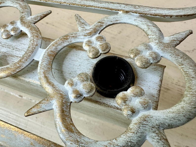 May include: Close-up of a decorative metal fixture with a distressed white and gold finish. The design features floral and curved elements, with a black socket in the center. The fixture is likely for lighting.