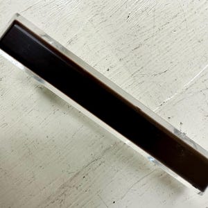 May include: A clear acrylic drawer pull with a black insert. The pull is rectangular in shape and has a smooth, polished finish.
