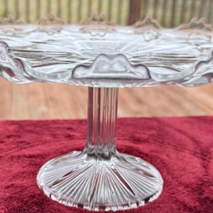 May include: A clear glass cake stand featuring a circular plate with a scalloped, raised edge and a pressed pattern. The stand is supported by a central, vertically ribbed pedestal base that flares out into a circular, rayed foot.