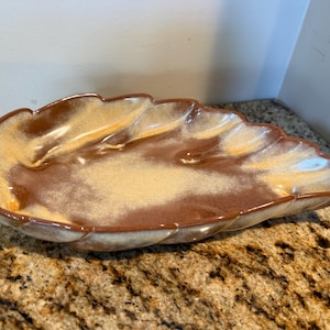 May include: A ceramic leaf-shaped serving dish featuring a glossy glaze with a blend of deep brown and creamy beige tones. The dish has a scalloped edge that mimics the natural contours of a leaf, with a smooth, reflective surface finish.