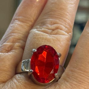May include: Silver-toned ring featuring a large, faceted, oval-shaped red gemstone. The gemstone is held in place by four prongs. The ring is shown on a finger.