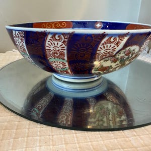May include: A decorative ceramic bowl with a blue rim and a white base. The bowl features a detailed pattern of red, white, and gold designs. The bowl is placed on a mirrored surface.