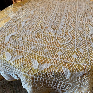 May include: A cream-colored, crocheted tablecloth with a delicate, openwork design. The intricate pattern covers a yellow table, creating a vintage aesthetic. The tablecloth's edges are scalloped, adding a touch of elegance to the overall look.