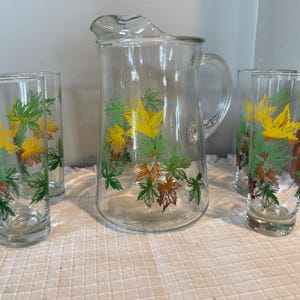 May include: A clear glass pitcher and four matching glasses. Each piece is decorated with a colorful leaf design in yellow, green, and brown. The pitcher has a handle and a pouring spout, perfect for serving beverages.