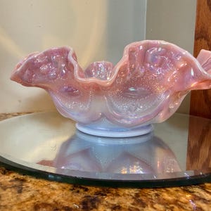 May include: A pale pink iridescent glass bowl with a ruffled, scalloped edge and a textured, embossed pattern featuring small raised dots and curved lines. The bowl has a circular base and displays a reflective, pearlescent finish throughout.