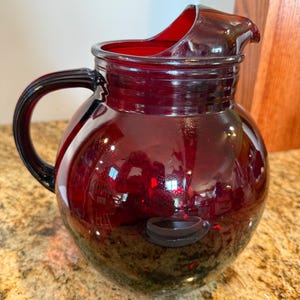 May include: A deep red glass pitcher with a rounded body, a wide pouring spout, and a curved handle. The glass is translucent and smooth, featuring a circular indentation on the side of the body and a ribbed neck design near the top rim.