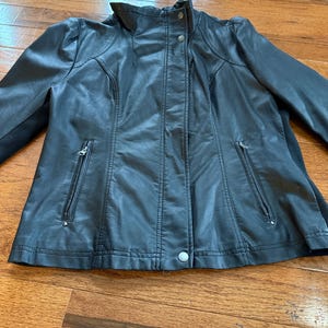 May include: A black faux leather jacket with a high collar and a fitted silhouette. The jacket features two zippered pockets, a button closure at the waist, and decorative stitching details. The jacket is displayed on a wooden surface.