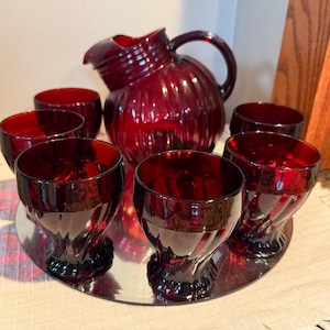 May include: A ruby red glass drinkware set, including a pitcher and six glasses, arranged on a mirrored tray. The pitcher has a handle and a ribbed design. The glasses have a twisted stem design.