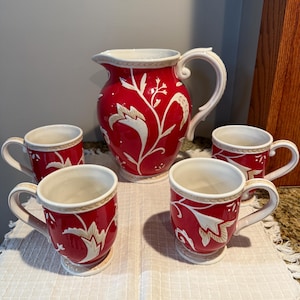 May include: A red ceramic pitcher and four matching mugs. The pitcher has a large handle and a white floral design. The mugs also have the same design. The set is on a white fringed cloth.