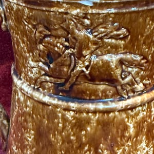 May include: Close-up of a brown ceramic pitcher with a detailed relief of a horse and rider. The pitcher has a textured surface and a dark brown rim. The handle is visible on the left side. The overall design suggests a rustic or antique aesthetic.