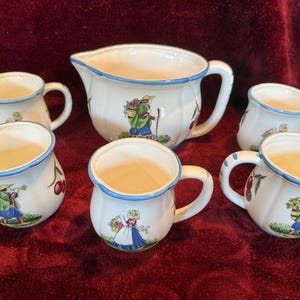 May include: A white ceramic pitcher and mugs set with a blue rim and handles. Each piece features a painted design of a person, cherries, and a floral motif. The set includes a pitcher and five mugs.