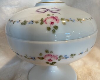 May include: White milk glass candy dish with a lid. The dish has a pedestal base and is decorated with a floral design of pink roses, blue flowers, and green leaves. The lid has a purple ribbon and a gold trim.