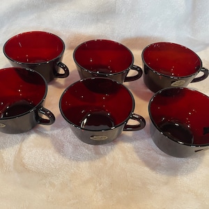 May include: Six red glass teacups with black rims and gold accents. The cups are arranged in a semi-circle on a white surface.