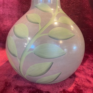 Puede incluir: Un jarrón redondo de cerámica gris con un diseño de hojas pintado en verde. El jarrón tiene una boca ancha y un fondo redondeado.