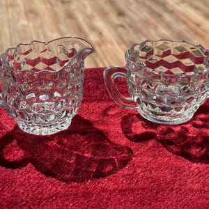 May include: A clear glass creamer and a matching sugar bowl featuring a raised geometric cube pattern. The creamer has a single handle and a pouring spout, while the sugar bowl has two handles on opposite sides. Both pieces have scalloped rims.
