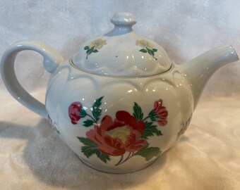 May include: White ceramic teapot with a floral design. The teapot has a rounded body with a scalloped edge and a lid with a knob. The floral design features red, yellow, and green flowers.