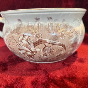 May include: Antique white and brown ceramic chamber pot with a handle. The pot features a floral design with tulips and small flowers. The rim has a gold trim. The pot is sitting on a red surface.