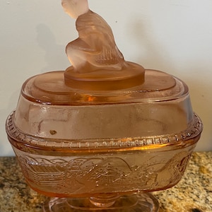 May include: A pink glass candy dish with a raised design of a cabin and trees. The lid features a figurine of a woman in a dress.
