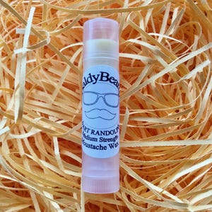 Może przedstawiać: Przezroczysty tubus z woskiem do wąsów z białą etykietą, na której widnieje napis "Baldy Beard" i "Bart Randolph Medium Strength Moustache Wax". Etykieta zawiera również kreskówkową ilustrację wąsów i okularów.