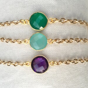 May include: Three gold chain bracelets with different colored gemstone charms. The top bracelet has a green gemstone, the middle bracelet has a light blue gemstone, and the bottom bracelet has a purple gemstone.