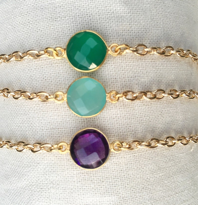 May include: Three gold chain bracelets with different colored gemstone charms. The top bracelet has a green gemstone, the middle bracelet has a light blue gemstone, and the bottom bracelet has a purple gemstone.