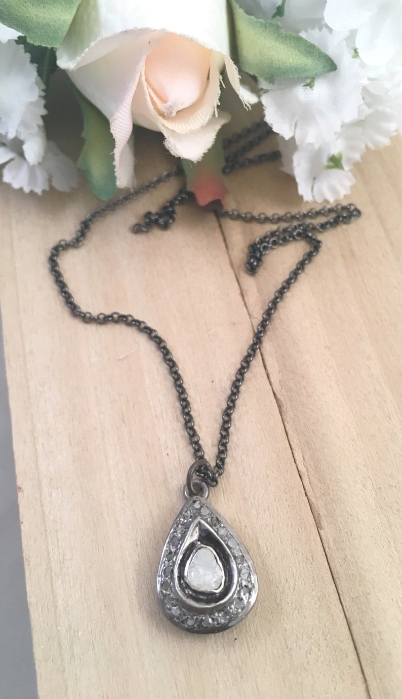 Puede incluir: Un collar con un colgante de plata en forma de l&aacute;grima, adornado con un gran diamante bruto claro en el centro. El colgante est&aacute; rodeado de diamantes m&aacute;s peque&ntilde;os y cuelga de una delicada cadena de plata.