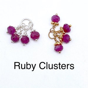 以下が含まれることがあります： 銀または金のチェーンとネックレスやブレスレットに取り付けるための小さなループが付いた、ルビーレッドの宝石のクラスターが2つ。クラスターは白い背景の上にあります。ルビーのクラスター