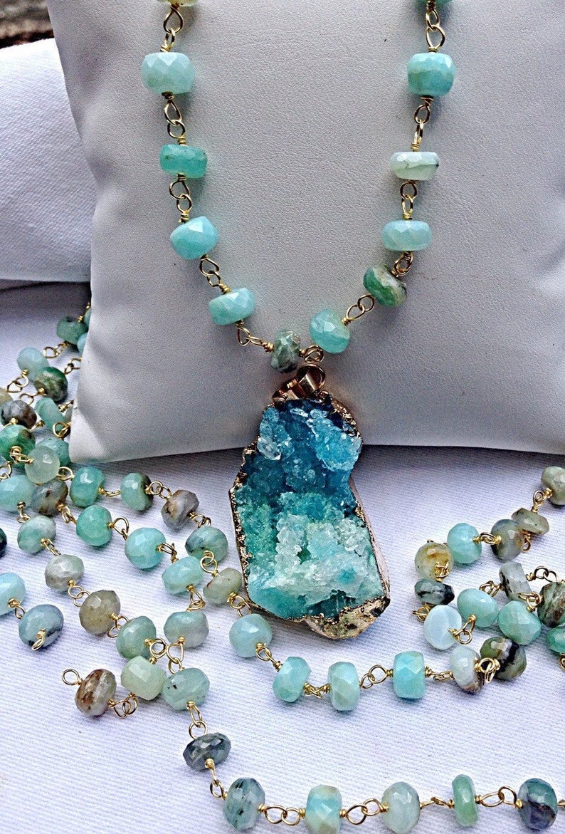 May include: A long, layered necklace with a large, aqua blue and green crystal pendant. The necklace is made of gold chain and small, faceted aqua blue beads.