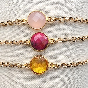 May include: Three gold chain bracelets with different colored gemstone charms. The top bracelet has a light pink charm, the middle bracelet has a pink and red charm, and the bottom bracelet has a yellow charm.