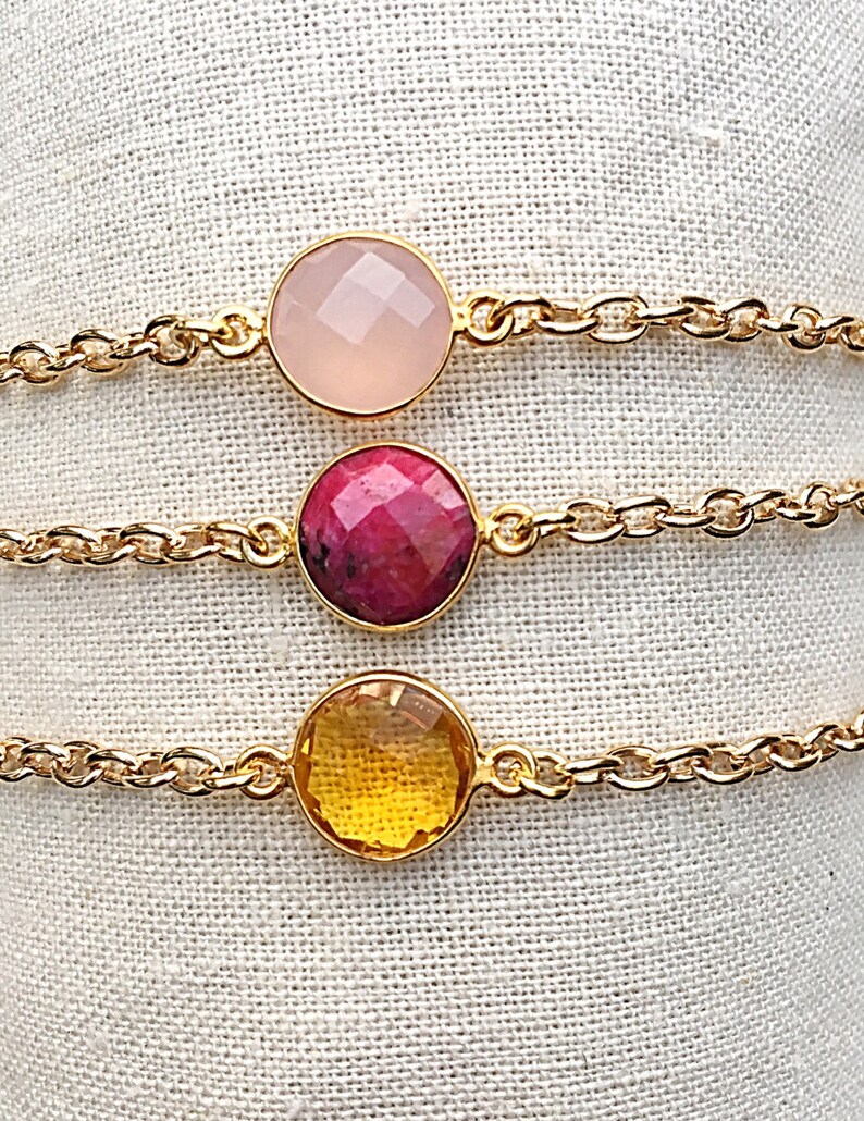 May include: Three gold chain bracelets with different colored gemstone charms. The top bracelet has a light pink charm, the middle bracelet has a pink and red charm, and the bottom bracelet has a yellow charm.