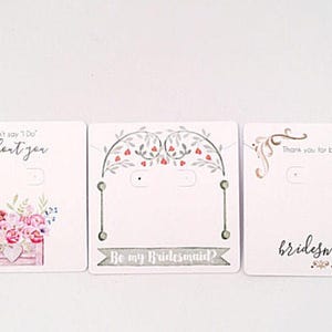 May include: Three white card earring displays with floral designs and text. The first card says "I can't say 'I Do' without you". The second card says "Be my Bridesmaid?" and the third card says "Thank you for being my bridesmaid."