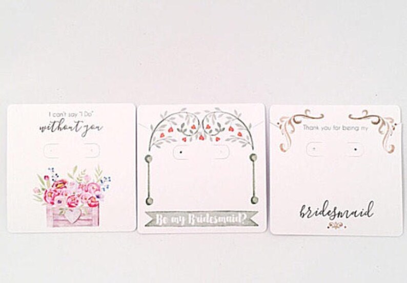May include: Three white card earring displays with floral designs and text. The first card says "I can't say 'I Do' without you". The second card says "Be my Bridesmaid?" and the third card says "Thank you for being my bridesmaid."