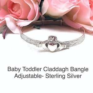 May include: A silver Claddagh bangle bracelet with an engraved design. The bracelet is adjustable and is shown on a white background with pink flowers.