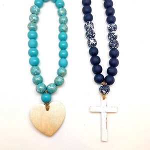 May include: Two beaded garlands with wooden charms. One garland is turquoise blue with a gold heart charm. The other garland is black with white speckles and a white wooden cross charm.