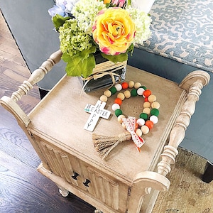 Può includere: Un tavolino da salotto in legno bianco con una croce fatta di piastrelle di legno che recita "Happy Easter" e una ghirlanda di perline con perline arancioni, bianche e verdi. Un vaso con un mazzo di fiori gialli, bianchi e blu si trova sul tavolo.