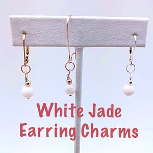 May include: Three pairs of white jade earring charms, each with a different metal finish: gold, rose gold, and silver. The charms are displayed on a white stand. White Jade Earring Charms.