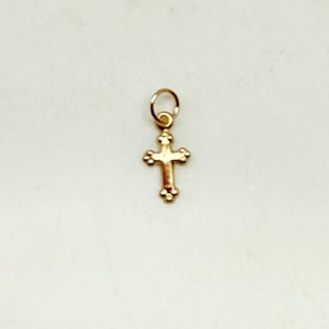 May include: A small, gold-colored cross pendant with a loop at the top for hanging. The cross has a detailed design with rounded edges and small decorative elements. The pendant is likely made of gold or a gold-toned metal.